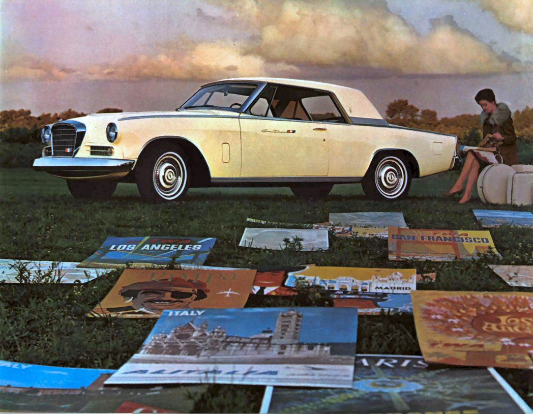 Brochure photo of a cream 1963 Gran Turismo Hawk on grass among travel posters, a woman seated at right