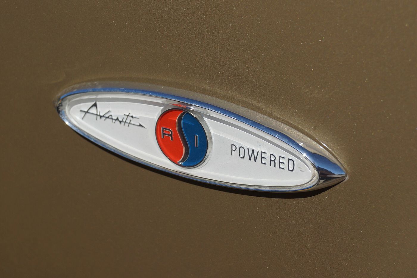 Close-up of an oval chrome and white badge reading Avanti, a red and blue R1 roundel, and Powered, on gold paint